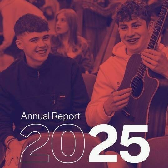 Two young musicians sitting and smiling with one holding guitar. Photo has red filter and text stating 'Annual Report 2025'.