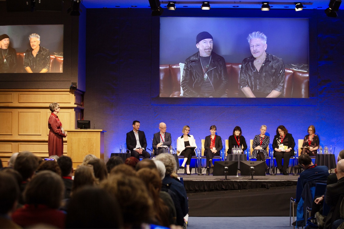 8 panellists sitting on stage with chair standing to right under screen displaying 2 musicians